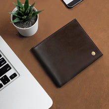 Load image into Gallery viewer, A smooth brown leather wallet with a small golden logo stud on a brown background next to a laptop.