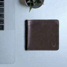 Load image into Gallery viewer, A closed brown leather bi-fold wallet for men rests beside a silver laptop and a small fresh plant.