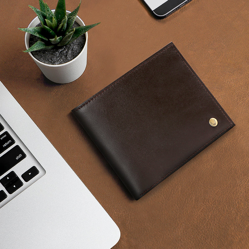 A smooth brown leather wallet with a small golden logo stud on a brown background next to a laptop.