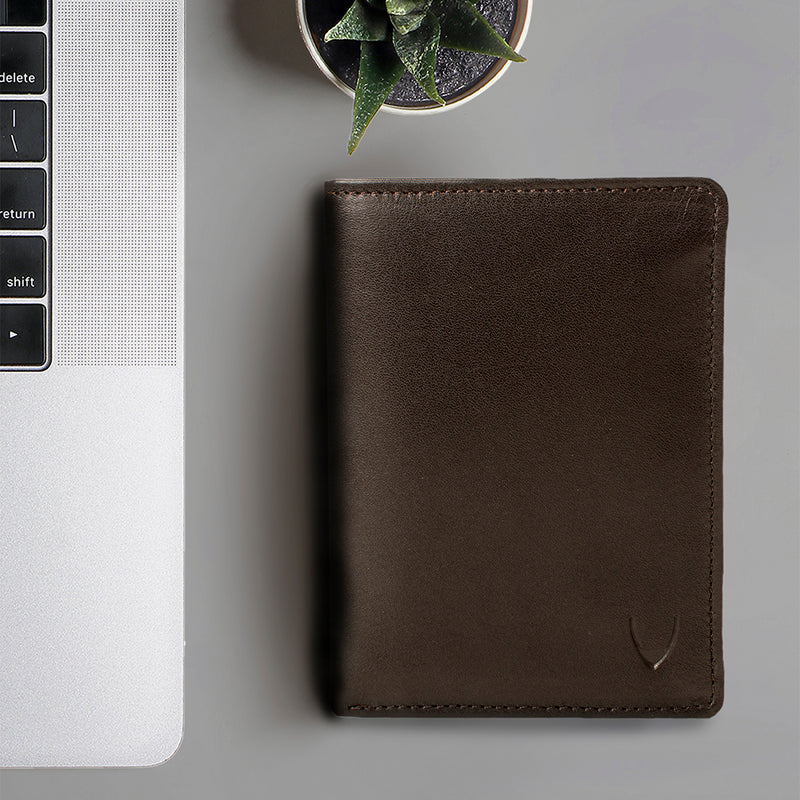 A closed dark brown bi-fold wallet resting beside a silver laptop & a small plant in the background.