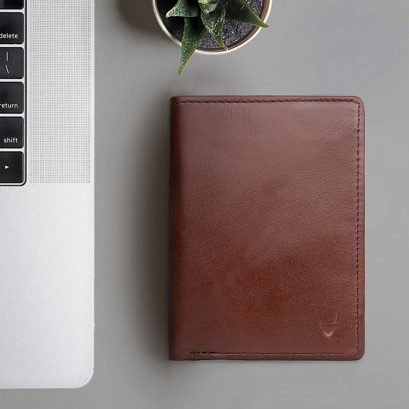A dark tan leather bifold wallet next to a silver laptop and a small succulent plant from Hidesign.