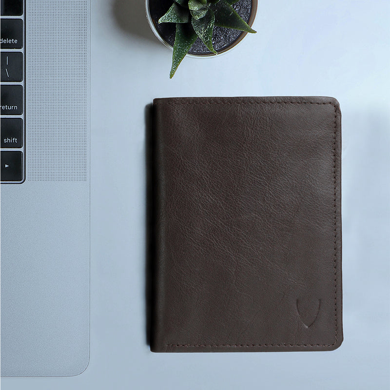 A brown men’s leather bi-fold wallet is placed on the desk beside a laptop and a small potted plant.